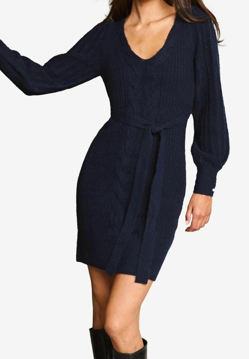 Marinblå stickad klänning med V-ringning, ribbad textur och långa puffärmar. Har ett tygbälte vid midjan och kabelstickat mönster.
