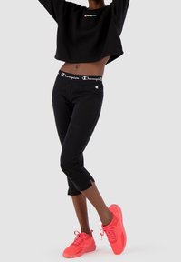 Felpa nera cropped con un piccolo logo colorato, abbinata a leggings neri alla caviglia con vita elasticizzata e scarpe da ginnastica rosa fluorescente.