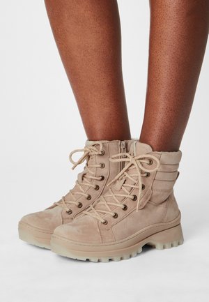 Lyse beige ruskind snørestøvler med polstrede kraver og tykke såler, båret på mørktonede ben mod en hvid baggrund.