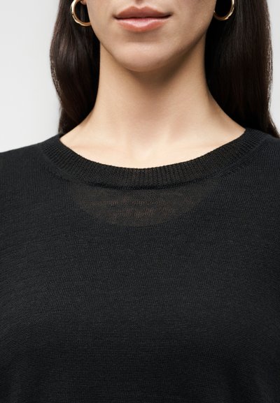 Κοντινό πλάνο μιας γυναίκας που φοράει χρυσούς κρίκους σκουλαρίκια και μαύρο πλεκτό πουλόβερ με στρογγυλή λαιμόκοψη, δείχνοντας τα χείλη, τον λαιμό και το πάνω μέρος του στήθους της.