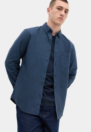 Jonge man met een blauw zwart geruit overhemd over een donkerblauw T-shirt met donkerblauwe spijkerbroek, staand met handen achter de rug.