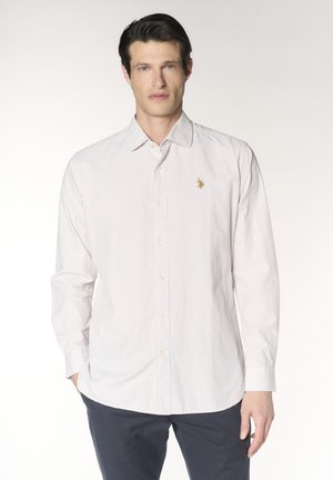 Uomo che indossa una camicia a righe rosa chiaro a maniche lunghe con piccolo emblema dorato, pantaloni scuri, in piedi davanti a uno sfondo semplice.