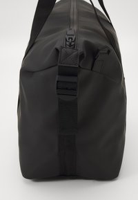 Sac de sport noir avec une texture lisse et mate, doté d'une fermeture éclair, d'une sangle ajustable et de poignées latérales. Design simple et rectangulaire.