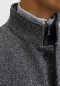 Hall hallivillase segumantlist mantel, millel on kõrge krae, lukuga sulgemine ja nupuga detail. Tekstuuriga kangas peene kalasaba mustriga.