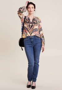 Bunte gemusterte Bluse mit langen Ärmeln, kombiniert mit blauen Slim-Fit-Jeans und schwarzen Lacklederschuhen. Eine kleine schwarze Schultertasche ist ebenfalls enthalten.