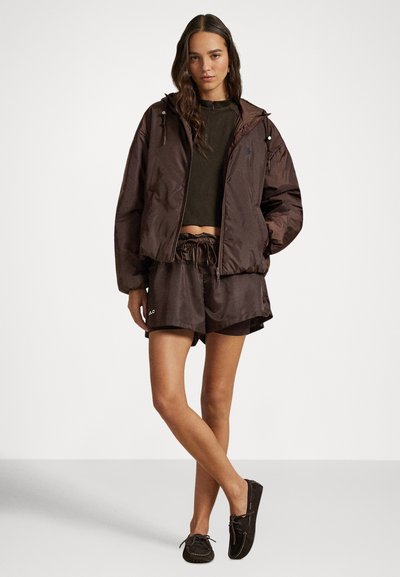 Καφέ κοντό μπουφάν με κουκούλα, συνδυασμένο με σκούρα σορτς και παπούτσια τύπου slip-on. Διαθέτει σκοινί στη μέση και διακριτική σήμανση.