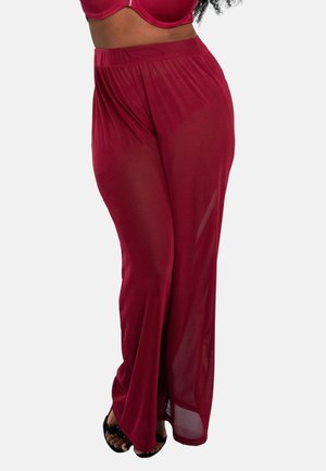 Frau trägt transparente bordeauxrote Hosen mit weitem Bein und passenden bordeauxroten BH, steht mit überschlagenen Beinen, barfuß in schwarzen Sandalen.