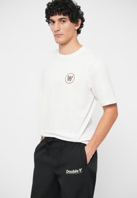 Bijela pamučna majica kratkih rukava s smešnim kružnim logotipom. U kombinaciji s crnim trenirkom koji ima bijeli otisnuti logotip.