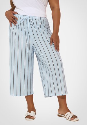 Kobieta ubrana w jasnoniebieskie, szerokie, krótkie spodnie z pionowymi czarno-białymi paskami oraz białe klapki.