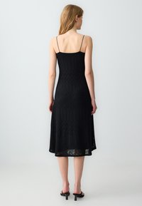 Robe noire, à mi-mollet, avec un buste ajusté et de fines bretelles, présentant un motif chevron texturé et un tissu léger et respirant.