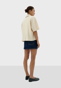 Beige short-sleeve shirt with a loose fit, paired with a dark denim skirt. The model wears black flat shoes and has a bun hairstyle.