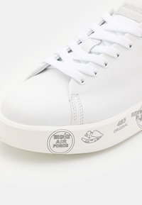 Gros plan sur une basket blanche montrant des lacets blancs, une tige en cuir lisse et une semelle imprimée avec l'inscription "White Air Force" et des motifs graphiques.