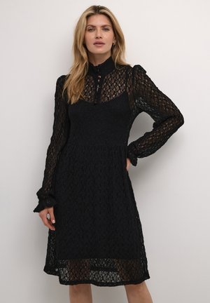 Robe en dentelle noire avec manches longues et col haut. Présente un motif texturé, une ouverture en forme de clé au niveau du cou, et des poignets de manches ondulés.