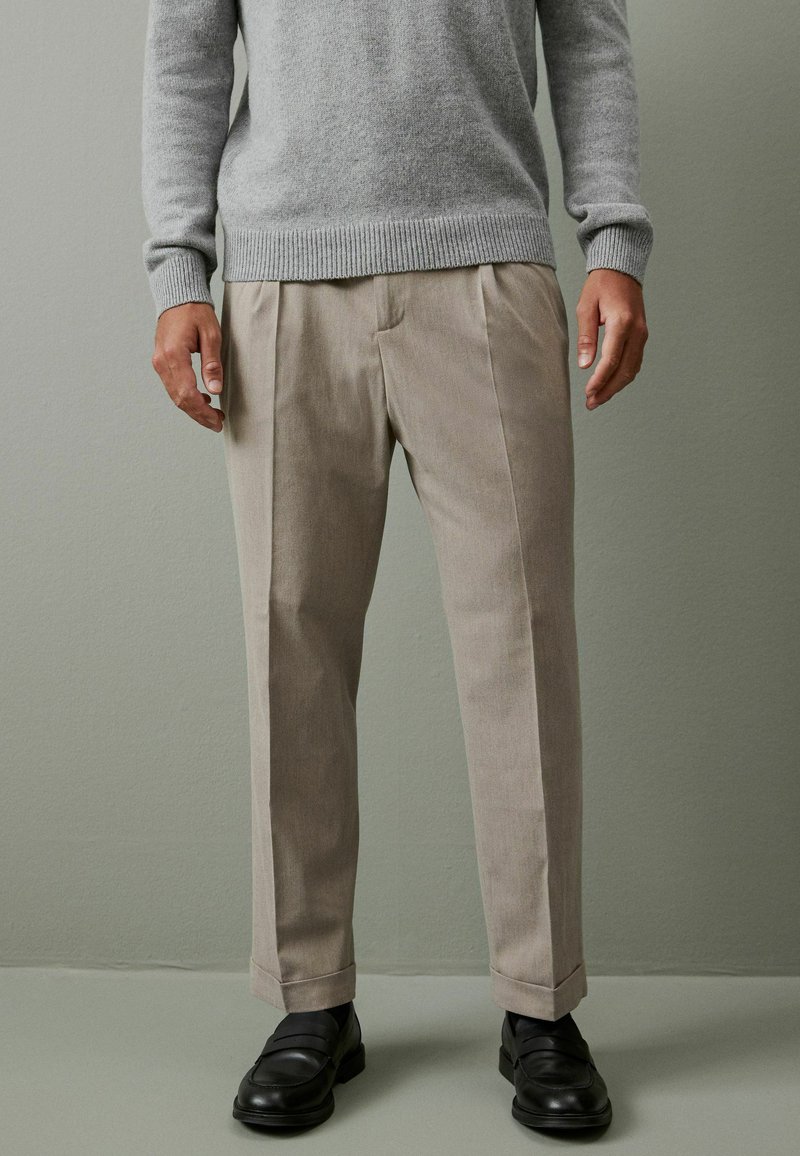 Hellgrauer Pullover mit gerippten Bündchen, kombiniert mit beigefarbenen, plissierten Hosen in taillierter Passform und schwarzen Loafers. Glatte Textur, lässiger Stil.