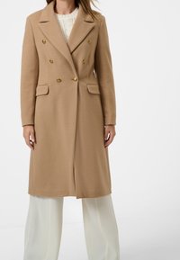 Abrigo beige de doble botonadura con botones dorados, bolsillos laterales y un ajuste entallado. La tela parece texturizada, complementada con pantalones blancos de pierna ancha.