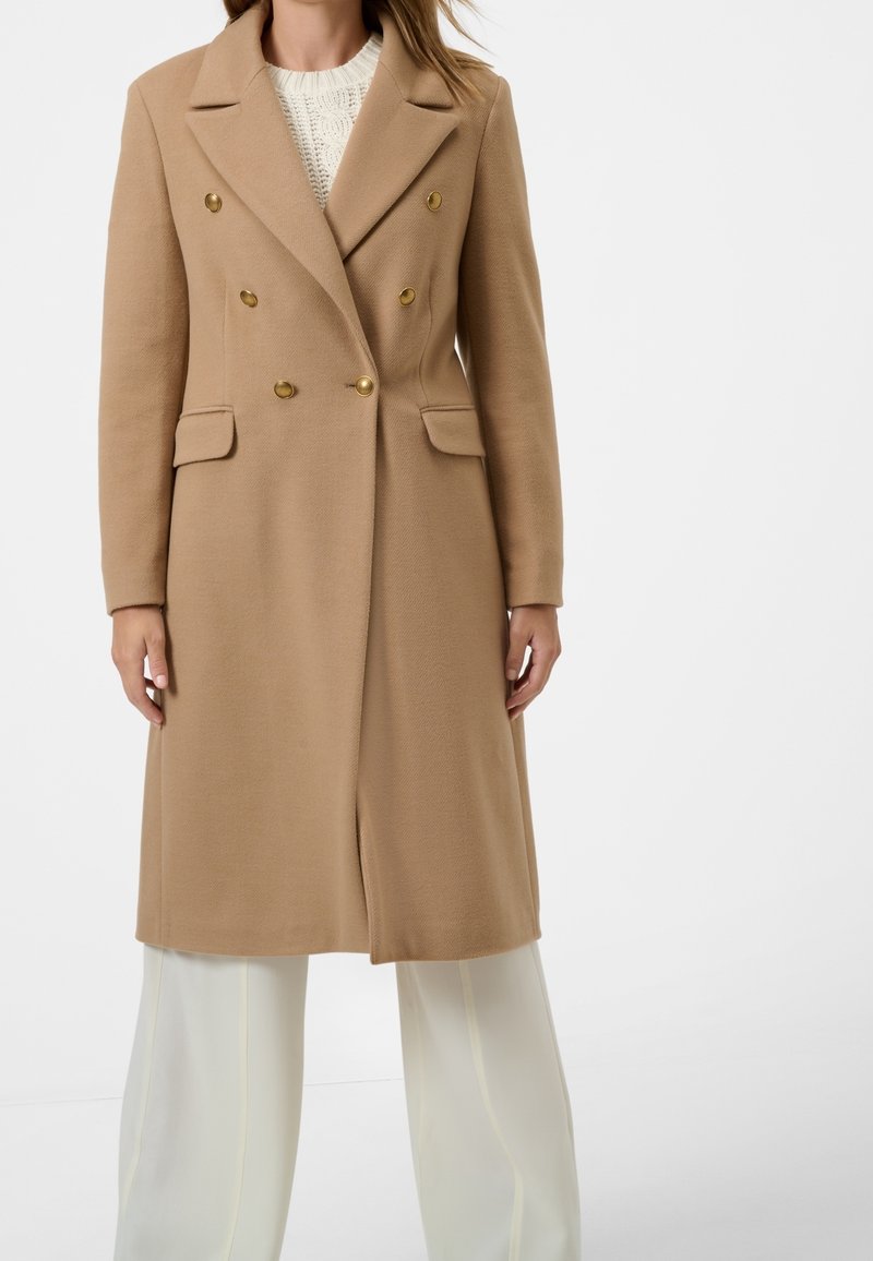 Abrigo beige de doble botonadura con botones dorados, bolsillos laterales y un ajuste entallado. La tela parece texturizada, complementada con pantalones blancos de pierna ancha.