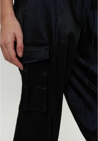 Černé cargo kalhoty vyrobené z hladké, lesklé tkaniny. Mají boční kapsu a design s uvolněným střihem. Zaměřte se na texturu a detail materiálu.