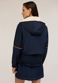 Giacca blu navy con colletto e cappuccio crema, con polsini elasticizzati e dettagli a striscia marrone sulle maniche, realizzata in tessuto leggero.