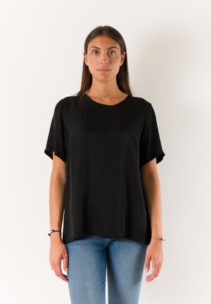 Donna con lunghi capelli scuri che indossa una maglietta nera semplice a maniche corte e jeans blu, in piedi davanti a uno sfondo bianco.