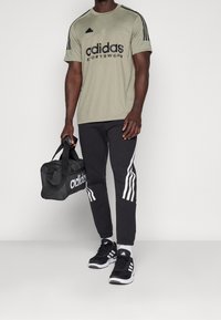 Herroutfit med en ljus olivgrön Adidas-tröja med svart logga, svarta joggers med vita ränder och svarta sportskor.