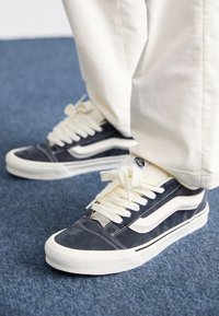 Zapatillas bajas de ante azul marino con detalles en cuero blanco, con cordones blancos y suelas de goma, sobre un fondo de moqueta azul.