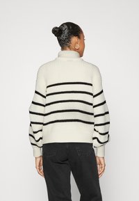 Le pull à col roulé en maille présente des rayures noires horizontales sur un fond crème. Les manches amples et les poignets côtelés ajoutent de la texture et de la forme.