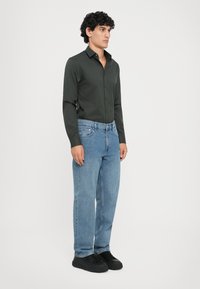 Camisa de botones de manga larga verde oscura combinada con vaqueros rectos de pierna clara y zapatillas negras. Diseño sencillo, ajuste estándar.