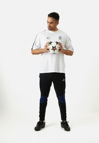 T-shirt de sport blanc avec logo, pantalon de survêtement noir et bleu, et crampons de foot noirs. Tient un ballon de football noir et blanc avec des accents verts.