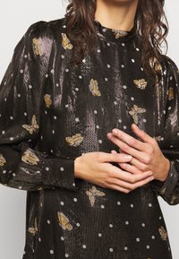 Femme portant un chemisier noir brillant avec des pois pâles et des motifs de papillons jaunes, mains doucement jointes au niveau de la taille.