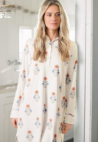 Vrouw met lang blond haar staat binnen en draagt een lichte lange mouw jurk met een bloemenpatroon van oranje en blauwe bloemen.