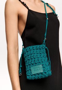 Borsa a tracolla in crochet di colore teal, con forma rettangolare, tracolla regolabile e superficie testurizzata, dotata di un'etichetta con il marchio sul davanti.