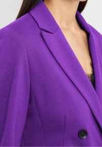 Žena v purpurovém blazeru s černým knoflíkem a viditelným černým krajkovým topem pod ním, zobrazená od ramene po polovinu hrudníku.