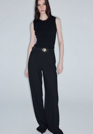 Vrouw met steil donker haar, gekleed in een mouwloos zwart topje, zwart broek met hoge taille en een zwarte riem, staand tegen een effen grijze achtergrond.