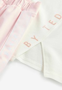 Roza i bijela kratka majica s kratkim rukavima s nabranim strukom, na kojoj je u svijetlo rozoj boji ispisano "BY TED BAKER" na bijelom platnu.