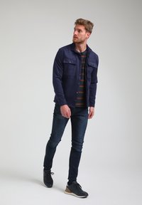 Giacca in camoscio blu navy con due tasche sul petto, abbinata a una camicia a righe, jeans slim e sneaker nere con un'accento beige.