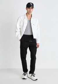 Weiße Denimjacke, graues, figurbetontes T-Shirt, schwarze Cargo-Hosen und weiße Sneaker mit schwarzen Akzenten. Schwarze Kappe mit Logo.