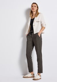 Blazer beige clair avec des revers structurés et un bouton, associé à un pantalon de jogging gris foncé avec des poches latérales et un cordon de serrage.