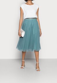 Vaaleansininen hame, jossa on poimutettu muotoilu, yhdistettynä valkoiseen paljeteilla koristeltuun toppiin. Mukana valkoinen laukku ja remmisandaalit.