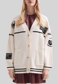 Kobieta w kremowym kardiganie w czarne paski i łatki, nosząca bordo sukienkę, stojąca na jednolitym szarym tle.