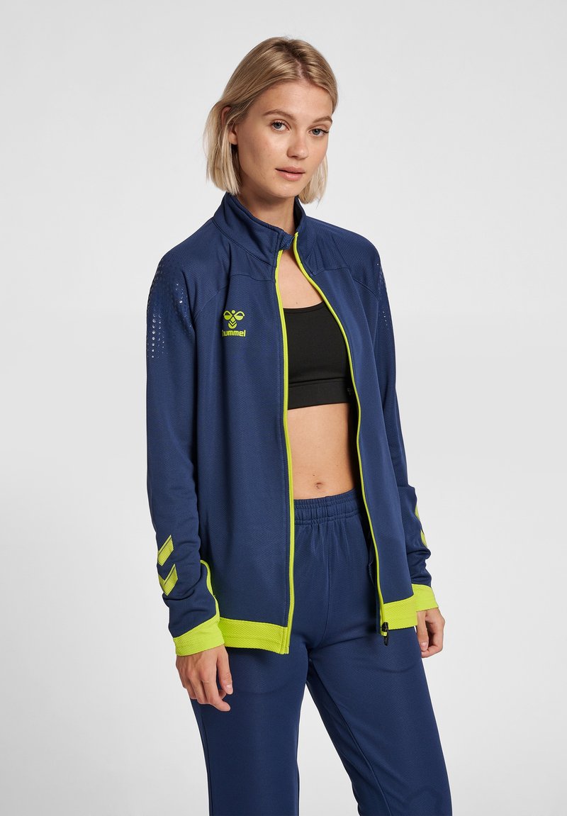Chaqueta navy con cremallera y acentos en verde lima, de tela texturizada y con detalles perforados en los hombros. Combinada con pantalones a juego.