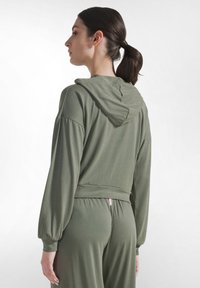 Kobieta z ciemnym kucykiem, ubrana w luźną oliwkowozieloną bluzę z kapturem i pasujące spodnie, widziana od tyłu na jednolitym tle.