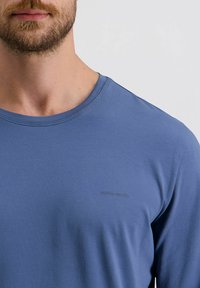 Bärtiger Mann, der ein blaues Rundhals-T-Shirt mit kleinem "Pierre Cardin"-Schriftzug auf der Brust trägt, vor einem einfarbigen Hintergrund.