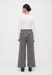 Femme aux cheveux foncés en chignon, portant un haut à manches longues blanc, un pantalon large à taille haute avec un imprimé zèbre noir et gris, et des bottes noires.