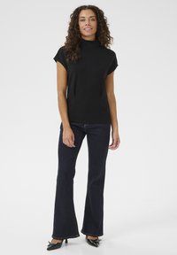 Melnas īsās piedurknes augsts kakls tops ar pieguļošu siluetu, apvienots ar tumši ziliem platiem džinsiem un melniem papēžiem, uz neitrāla fona.