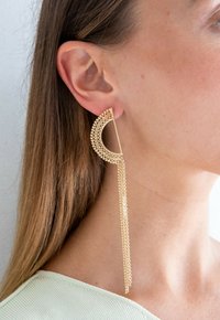 Gouden oorbellen in een halvemaanvorm met een gestructureerd patroon, bevestigd met een ketting die onder de oorlel hangt, met een gepolijste afwerking.