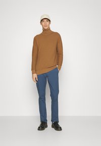 Maglione a collo alto a coste marrone, pantaloni blu e scarpe nere. Il modello indossa un berretto crema. Design semplice e aderente senza motivi visibili.