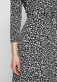 Czarna sukienka z białym wzorem w liście, z długimi rękawami, podkreśloną talią i zwiewnym dołem. Teksturowany materiał o subtelnym połysku.