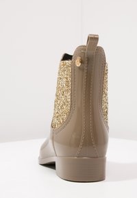 Botas de lluvia en color beige con un material brillante y resistente al agua, y un panel trasero cubierto de purpurina dorada; presentan un pequeño logotipo y una tira de agarre para ponérselas fácilmente.