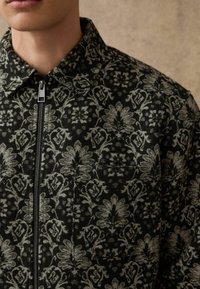 Schwarze und graue, mit Blumenmuster versehene Reißverschlussjacke aus strukturiertem Stoff, ausgestattet mit einem Kragen und einer Brusttasche.