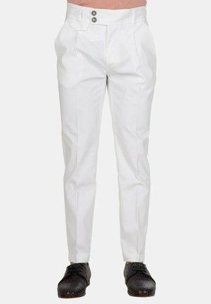 Pantaloni bianchi su misura con pieghe frontali e chiusura doppio bottone abbinati a scarpe nere testurizzate con lacci su sfondo chiaro.